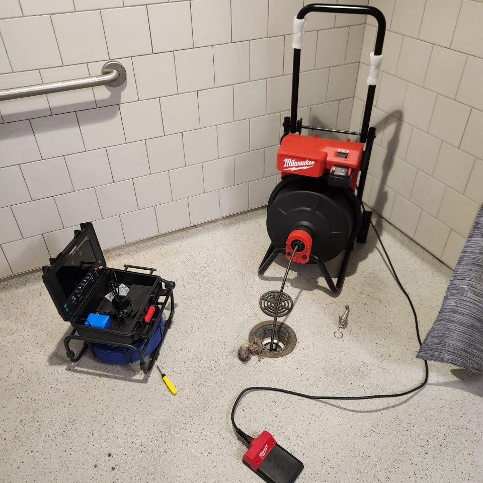Drain cleaning machine on a cart, feeding cable into an open floor drain, with a sewer camera case beside it